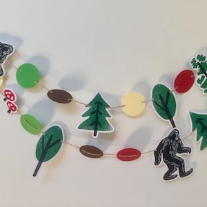 May include: A whimsical garland featuring cut-out shapes of trees, mushrooms, Bigfoot, and colorful circles. The garland is strung together with twine, creating a playful and decorative element. The color palette includes green, red, brown, and yellow.