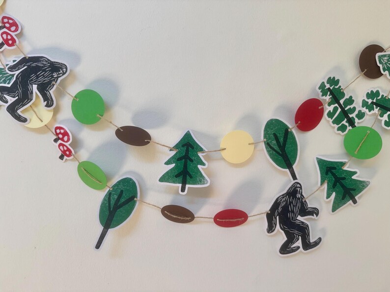May include: A whimsical garland featuring cut-out shapes of trees, mushrooms, Bigfoot, and colorful circles. The garland is strung together with twine, creating a playful and decorative element. The color palette includes green, red, brown, and yellow.