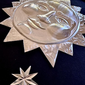 May include: A metallic silver sun wall art piece with a detailed face and textured rays, accompanied by two separate silver star accents. The entire set is mounted against a solid black background, creating a high-contrast display of celestial shapes.