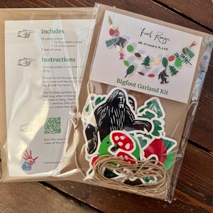 May include: A Bigfoot Garland Kit in a clear plastic package. The kit includes paper pieces, twine, and instructions. The package features a Bigfoot illustration, trees, and mushrooms. The text "Foot Range Mountain Bigfoot Garland Kit" is visible.