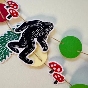 May include: A whimsical garland featuring a black Bigfoot silhouette, red mushrooms with white spots, green circles, and a green tree shape. The decorative elements are strung together with twine, creating a playful, nature-themed decoration.