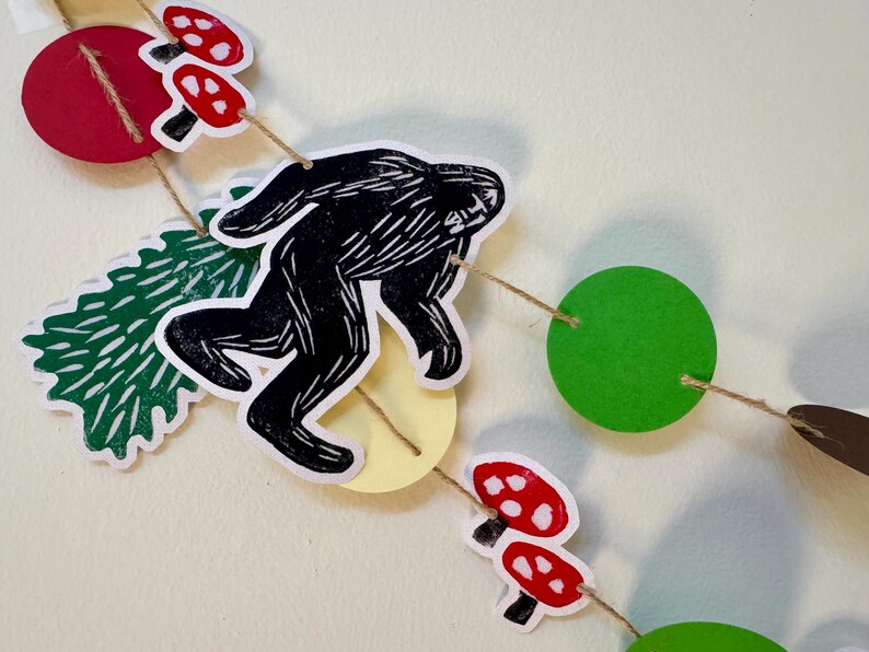 May include: A whimsical garland featuring a black Bigfoot silhouette, red mushrooms with white spots, green circles, and a green tree shape. The decorative elements are strung together with twine, creating a playful, nature-themed decoration.