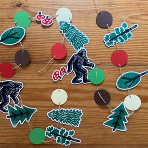 May include: A whimsical garland featuring cut-out shapes of Bigfoot, trees, leaves, mushrooms, and circles in green, red, brown, and cream colors. The shapes are strung together with twine, creating a playful forest theme.