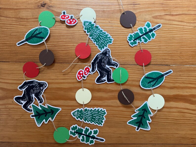 May include: A whimsical garland featuring cut-out shapes of Bigfoot, trees, leaves, mushrooms, and circles in green, red, brown, and cream colors. The shapes are strung together with twine, creating a playful forest theme.