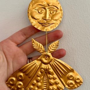 May include: Gold metal ornament featuring a moon with a face and a moth with intricate details. The ornament is suspended from a string.