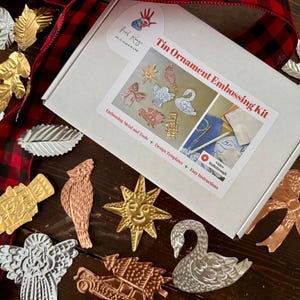 May include: A white box labeled "Tin Ornament Embossing Kit" with various embossed tin ornaments in gold, silver, and copper. Ornaments include a swan, angel, star, and bow. A red and black plaid ribbon and leaf ornaments are also present.