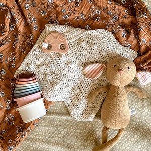 May include: A brown and white floral patterned swaddle blanket, a beige crocheted blanket, a brown stuffed bunny with pink ears, and a set of stacking cups in various colors are arranged on a beige and white patterned crib sheet.
