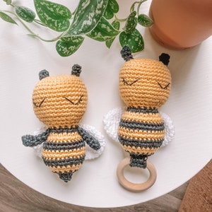 May include: Two crocheted bee toys with black and yellow stripes and white wings. One bee has a wooden ring attached to its body.