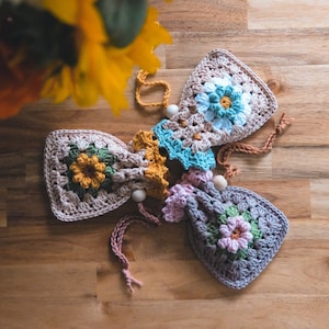 Könnte beinhalten: Drei gehäkelte Beutel mit Kordelzug und Blumendesigns in Rosa-, Blau- und Gelbtönen. Die Beutel sind im Granny-Square-Muster gearbeitet und haben Holzperlen an den Kordeln.