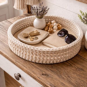 Könnte beinhalten: Ein runder, cremefarbener, gewebter Korb enthält eine kleine weiße Vase mit Lavendel, ein kleines Ledertablett mit Schlüsseln, ein kleines Notizbuch, Holzperlen und eine schwarze Sonnenbrille. Der Korb steht auf einem Holztisch.