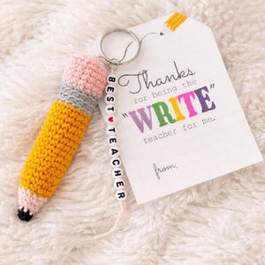 Puede incluir: Un llavero de lápiz de ganchillo hecho a mano con una goma de borrar rosa, una banda gris y un cuerpo amarillo. El llavero tiene una cadena de cuentas que dice "BEST TEACHER". Una etiqueta dice "Thanks for being the WRITE teacher for me."