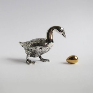 Op de afbeelding: Een gepolijst zilverkleurig ganzenbeeldje met gedetailleerde verentextuur op het lichaam en een gladde nek. De gans is gepositioneerd tegenover een klein, glad, goudkleurig eivormig object dat op het oppervlak rust.