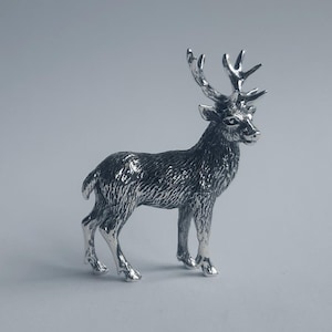 May include: A small, metallic silver-coloured figurine of a standing deer. The sculpture features detailed, textured fur across its body and a set of prominent, branching antlers on its head. The deer is depicted in a profile view, standing upright.