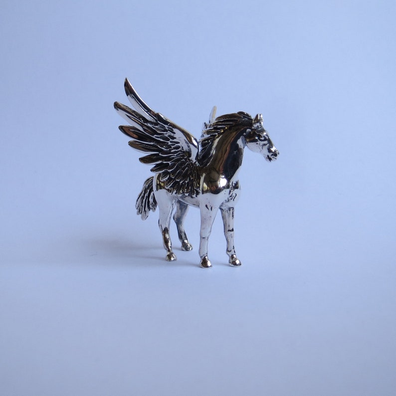 May include: A silver figurine of a winged horse, with a detailed sculpted body and wings. The horse is standing on a white background.
