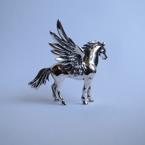 May include: A silver figurine of a winged horse, Pegasus, with detailed wings and a muscular body. The horse is standing on a white surface.