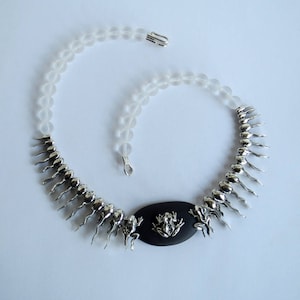 May include: A silver necklace with a black oval pendant featuring a frog design. The necklace is made with clear glass beads and silver frog charms.