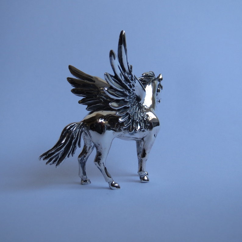 May include: A silver figurine of a winged horse with a detailed sculpted design. The horse is standing on all four legs with its wings spread out.