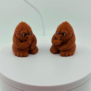 May include: Two identical brown, textured figurines shaped like small, furry creatures. Each figure has a rounded body, large eyes, and a hand raised to its mouth in a shushing gesture. The figures are seated and feature detailed fur patterns.