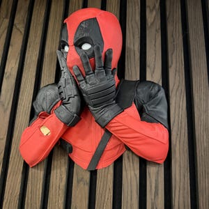 Puede incluir: Un busto de pared del personaje Deadpool, con una máscara roja y negra con lentes oculares blancos. La figura se muestra con ambas manos levantadas hacia el rostro, usando guantes tácticos negros y un traje rojo y negro con hombreras.