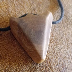 Könnte beinhalten: Ein Holzanhänger in Dreiecksform, an einer schwarzen Kordel befestigt. Der Anhänger hat eine glatte, polierte Oberfläche mit natürlichen Holzmaserungsmustern. Der Hintergrund ist eine strukturierte, hellbraune Oberfläche.