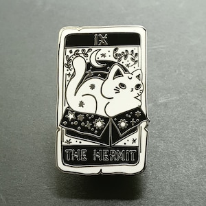 May include: A silver enamel pin featuring a white cat sitting in a box with stars. The box is open and the cat is looking to the right. The pin is a tarot card design and says "The Hermit" and "IX" on it.