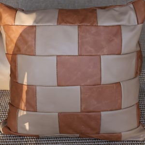 May include: A square cushion with a patchwork design. The cushion features alternating rectangular blocks of light tan and medium brown leather, stitched together. Ideal for home decor.
