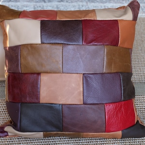 Peut inclure: Un coussin décoratif carré avec un motif patchwork. Le coussin est composé de carrés de cuir de différentes couleurs, dont beige, marron, bordeaux et rouge. Le coussin mesure environ 46 cm de côté.