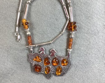 Peut inclure: Un collier en argent avec un pendentif représentant un motif d'oiseau stylisé et des pierres d'ambre. Le collier est composé de perles d'argent et de perles d'ambre.