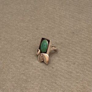 May include: A silver ring with a turquoise stone set in a rectangular frame. The ring has a decorative design with leaves and swirls.