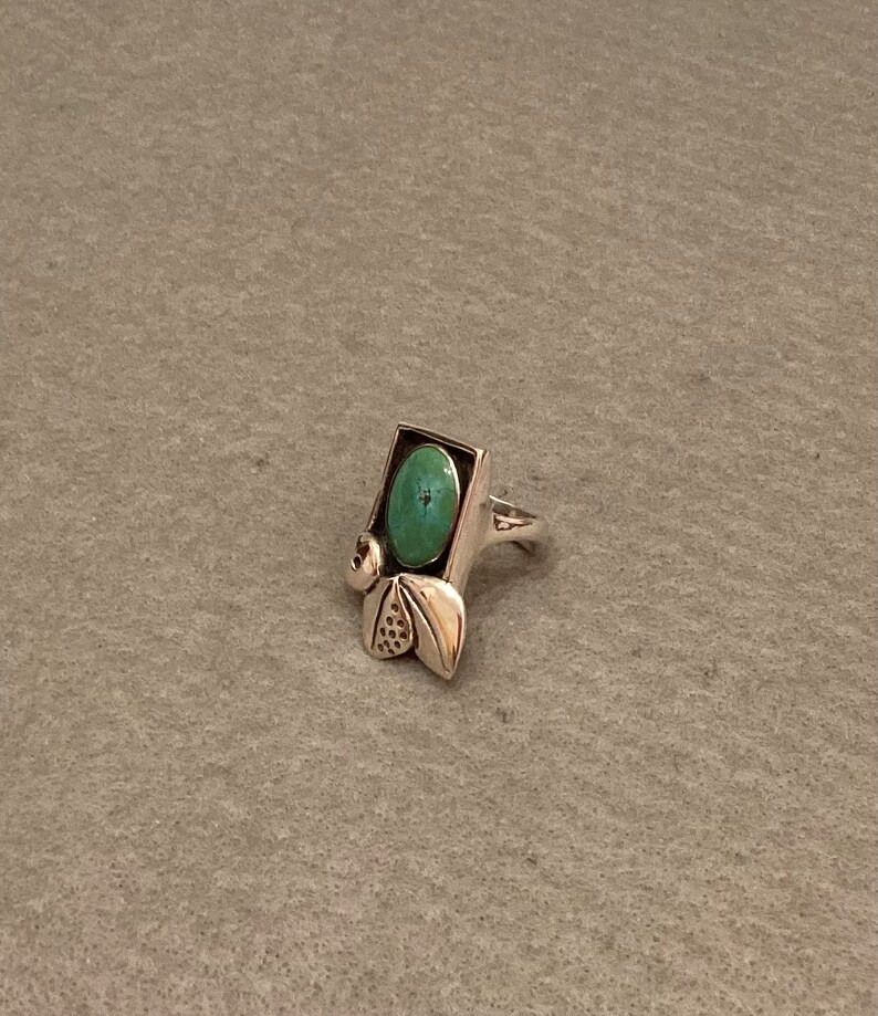 May include: A silver ring with a turquoise stone set in a rectangular frame. The ring has a decorative design with leaves and swirls.