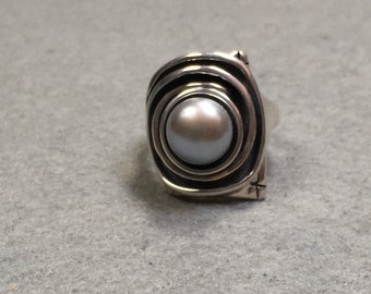 May include: A silver ring with a large, round, gray pearl set in a swirling, textured design.
