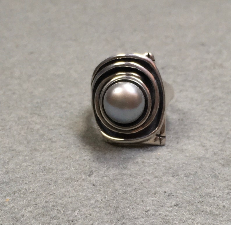 Peut inclure: Une bague en argent avec une grosse perle ronde grise sertie dans un motif tourbillonnant et textur&eacute;.