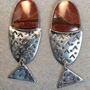 May include: A pair of silver earrings with a red and brown stone set in the top. The earrings are shaped like a fish with a triangular tail.