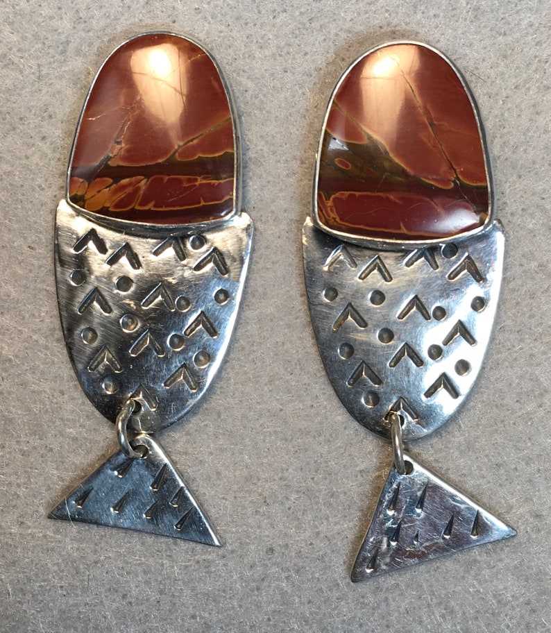 May include: A pair of silver earrings with a red and brown stone set in the top. The earrings are shaped like a fish with a triangular tail.