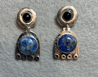 Peut inclure: Une paire de boucles d'oreilles pendantes en argent avec un haut circulaire orné d'une pierre noire et une base en forme d'arche. La base contient une pierre ronde en lapis-lazuli bleu sculptée d'un visage humain et quatre petites découpes.
