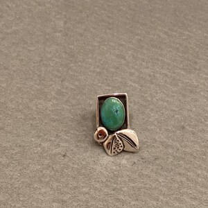May include: A silver pendant with a turquoise stone set in a geometric frame. The pendant features a stylized design with leaves and a small circle.