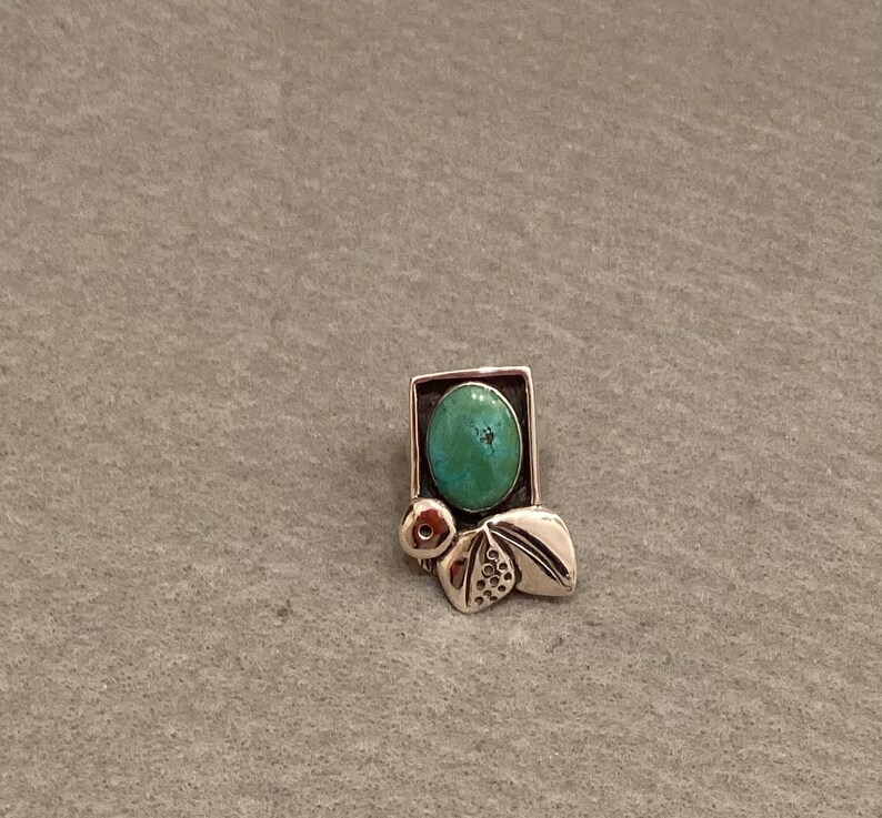 May include: A silver pendant with a turquoise stone set in a geometric frame. The pendant features a stylized design with leaves and a small circle.