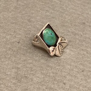 May include: A silver ring with a turquoise stone set in a square frame. The ring has a decorative design on the side.