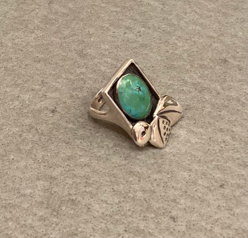 May include: A silver ring with a turquoise stone set in a square frame. The ring has a decorative design on the side.