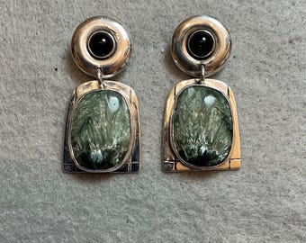 Peut inclure: Une paire de boucles d'oreilles pendantes avec un sommet circulaire en argent orné d'une pierre noire centrale, relié à une base rectangulaire en argent contenant une pierre vert mousse texturée au motif fibreux et rayonnant.