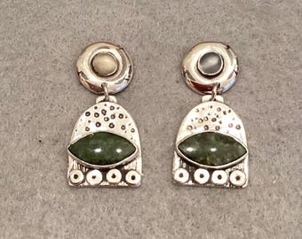 Peut inclure: Une paire de boucles d'oreilles argentées avec un haut circulaire orné d'une petite pierre, relié à une pièce pendante en forme d'arche. L'arche présente une pierre verte horizontale centrale, une section supérieure texturée et quatre petits cercles.