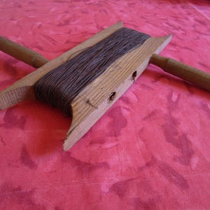 Op de afbeelding: Een vintage houten draadwinder met donkerbruin garen. De winder heeft een rechthoekig frame met twee houten handvatten. De draad is strak om het frame gewikkeld. De winder ligt op een roze ondergrond.