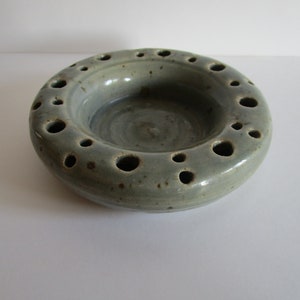 Puede incluir: Un plato de cerámica gris claro con un anillo de agujeros alrededor del borde. El plato tiene un esmalte moteado y un borde ligeramente elevado.