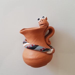 Puede incluir: Un jarrón de cerámica de terracota hecho a mano con una serpiente enrollada alrededor. La serpiente está pintada con rayas negras, blancas y azules. El jarrón tiene un asa y una pequeña cabeza de animal en la parte superior con ojos blancos.