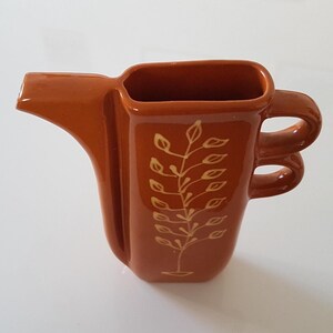 May include: A ceramic pitcher in a warm orange hue, featuring a unique design with a spout and a double-loop handle. The pitcher is adorned with a delicate, cream-colored botanical pattern, adding a touch of elegance to its form.
