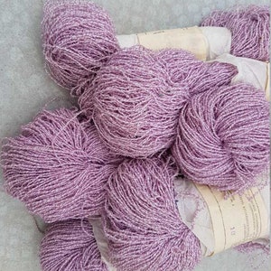 May include: A collection of lavender-colored yarn skeins, tightly wound into round shapes. The yarn has a textured appearance with a subtle sheen. Some skeins are partially wrapped in aged paper, adding a vintage aesthetic. The image showcases a craft supply, perfect for knitting or crochet projects.
