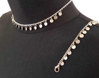 Puede incluir: Juego de collar y pulsera plateados. El collar es de estilo gargantilla con pequeños colgantes circulares. La pulsera refleja el diseño del collar, con colgantes similares y un cierre de broche. Las joyas se exhiben en un maniquí negro.
