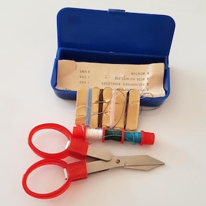 Puede incluir: Un pequeño kit de costura azul con tapa con bisagras, que contiene carretes de hilo, agujas y unas tijeras rojas pequeñas. El kit incluye tarjetas de hilo doradas y un inserto de papel con texto. Las tijeras tienen mangos rojos.