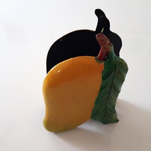 May include: A mango-shaped ornament with a glossy yellow finish. It has a green leaf and a black backing, creating a three-dimensional effect. The mango has a realistic appearance, suggesting a tropical theme.