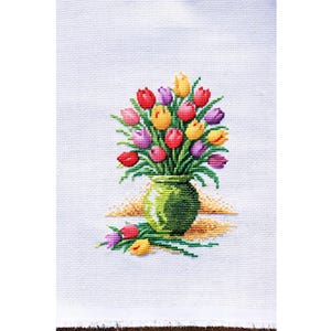 May include: Cross-stitch embroidery of a bouquet of tulips in a green vase. The tulips are red, yellow, and purple. The vase sits on a yellow and brown surface, with a few loose tulips nearby. The background is white.